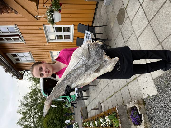 Waitress proudly holds a large dried stockfish outside a rustic wooden building