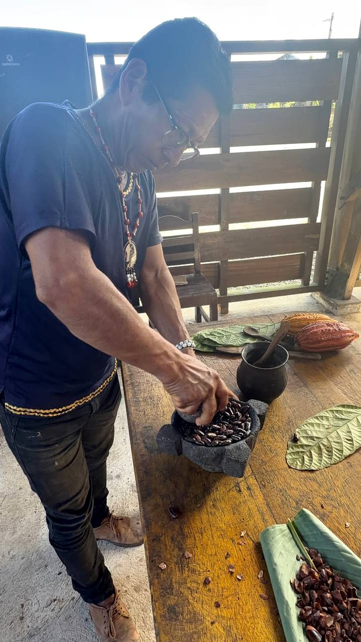 A person grinds roasted cacao beans on a stone mortar at a rustic wooden table, with cacao pods, leaves and a black cup beside them.