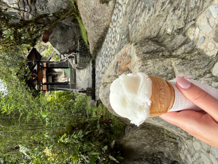 A hand holds a vanilla ice-cream cone in front of a cobbled pathway leading through lush greenery toward a small pavilion.