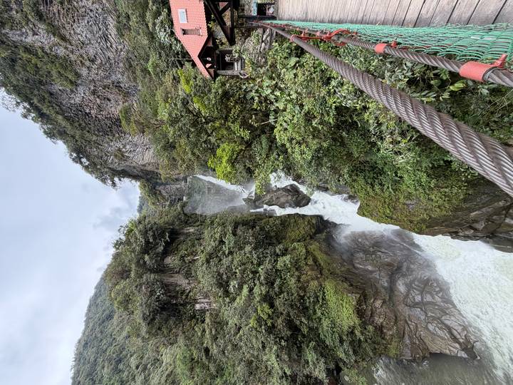 A powerful waterfall cascades through a lush green canyon viewed from a suspension bridge cable.