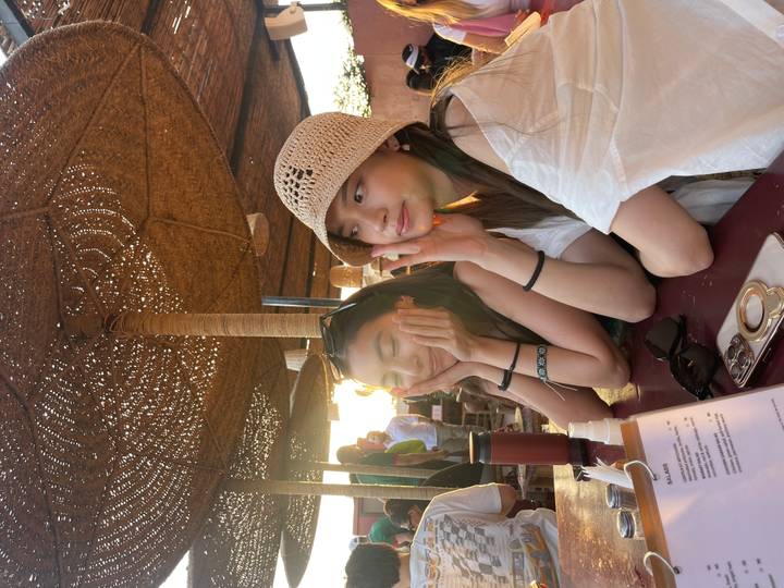 Two friends relax at a sunset rooftop café under woven parasols with warm backlighting.