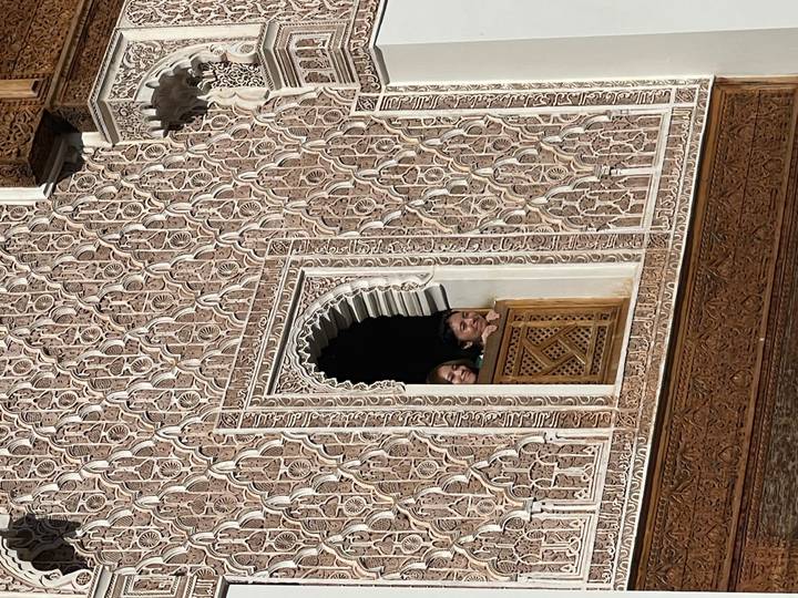 Two visitors peek out of an ornate Moorish window framed by intricate stucco carvings at Ben Youssef Madrasa.