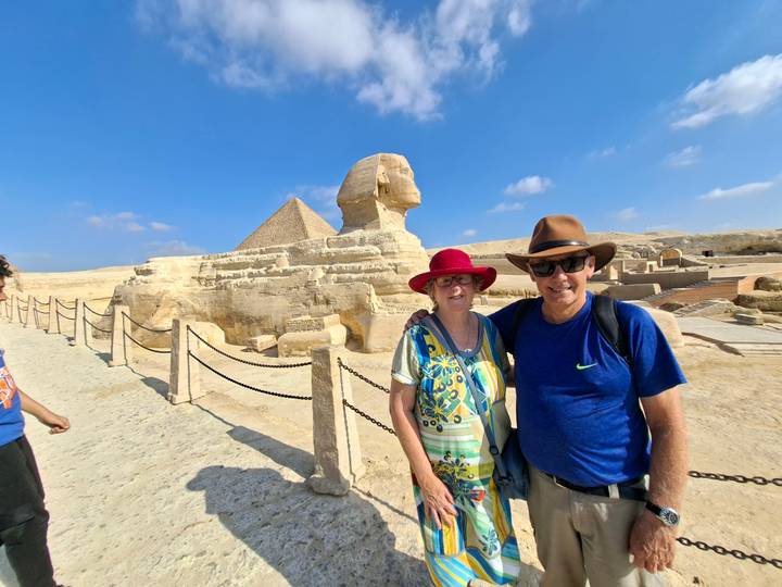 Couple standing before the Great Sphinx and a pyramid under blue skies.