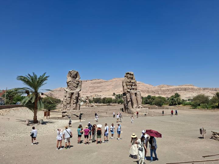 Tourists walk around the massive Colossi of Memnon statues under a clear blue sky.