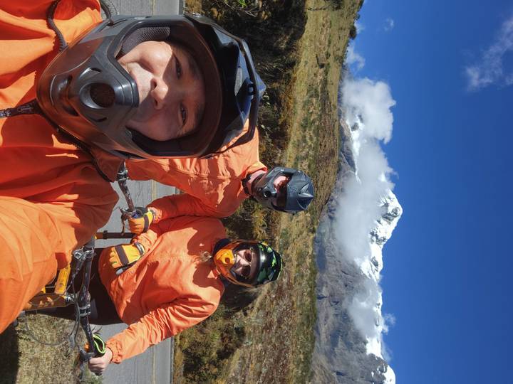 Three mountain bikers in orange jackets pose for a selfie with a snow-capped Andean peak behind.