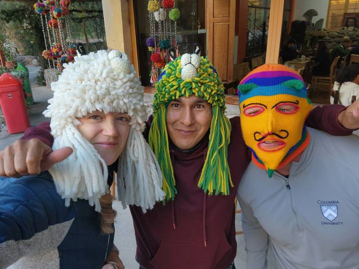 Three friends wear colorful traditional knit masks and smile at the camera in a busy market street.