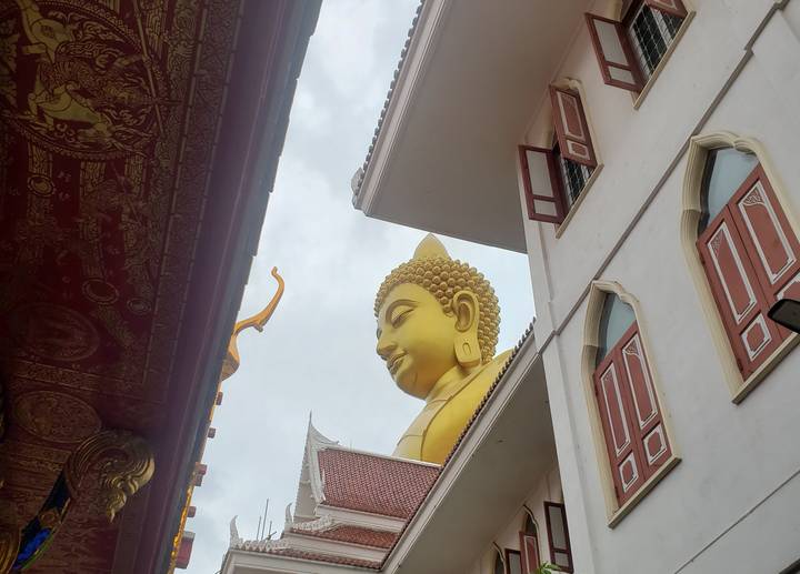 Upward perspective of a large golden Buddha head framed by temple buildings under a cloudy sky.