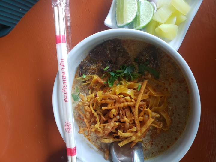 Close-up of a bowl of creamy noodle soup topped with crispy noodles and herbs, chopsticks resting above.