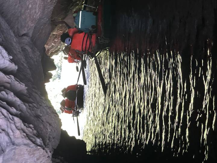 Rowboats of life-jacketed travelers paddle through a dim limestone cave toward daylight.