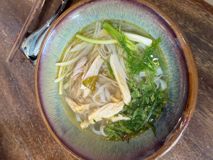 Bowl of Vietnamese pho soup with chicken strips, herbs, and rice noodles in clear broth.