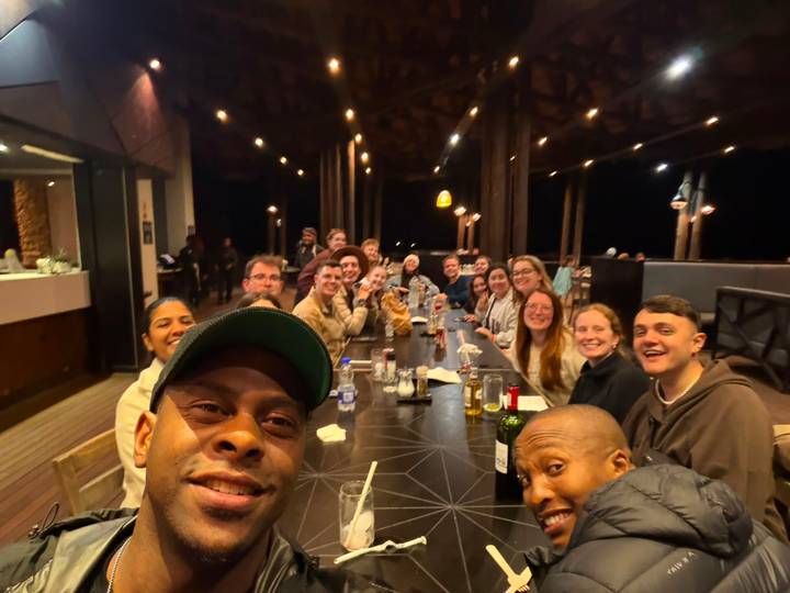 Large tour group enjoys lively dinner gathering at long table in open-air restaurant at night.
