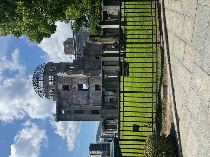 The ruined Atomic Bomb Dome rises behind an iron fence under a bright sky with patches of cloud and fresh green lawn in front.
