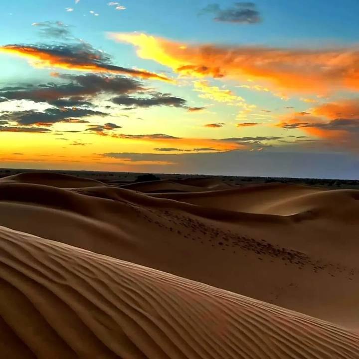 Rolling sand dunes glow under a colourful sunset sky streaked with orange, yellow and blue.