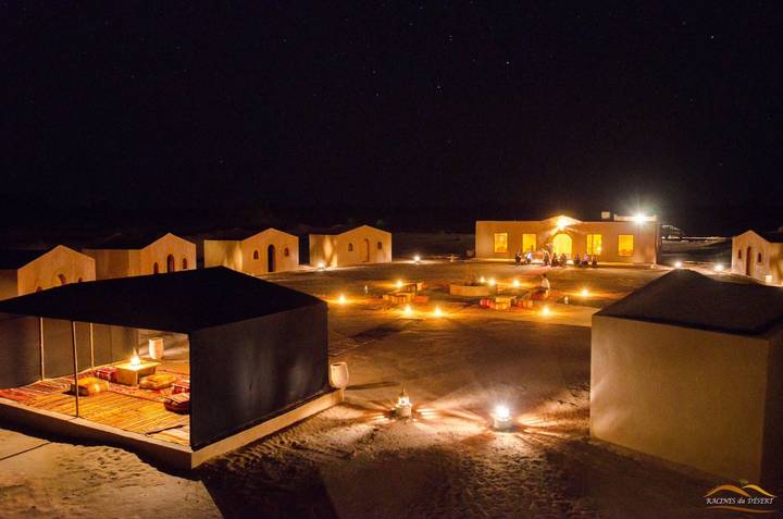 Stylish desert camp lit by dozens of lanterns and candles under a starry night sky; guests relax around low tables.