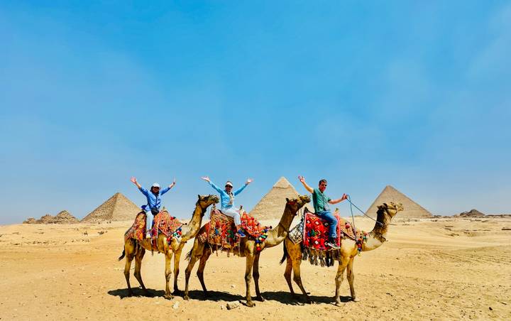 Riders on richly decorated camels raise their arms in front of the pyramids on the Giza Plateau under a blue sky.