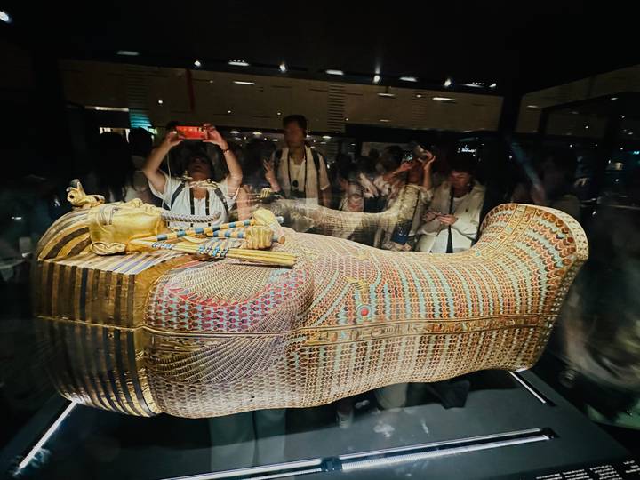 Golden sarcophagus exhibit behind glass attracts crowds of museum visitors taking photos and reflections.