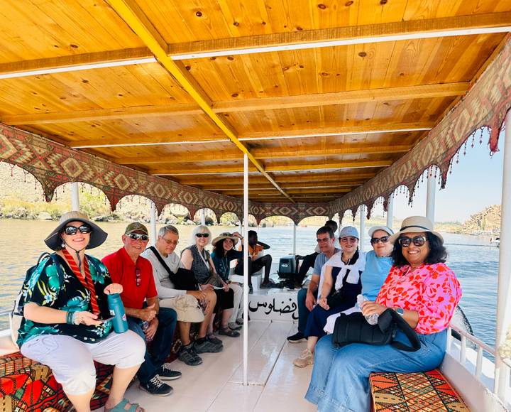 Tourists sit under a wooden roof on a traditional boat cruising the Nile, smiling toward the camera.
