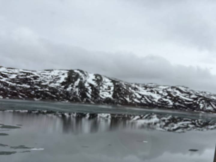 Blurry view of snow-covered mountains reflected in a still lake beneath grey clouds.