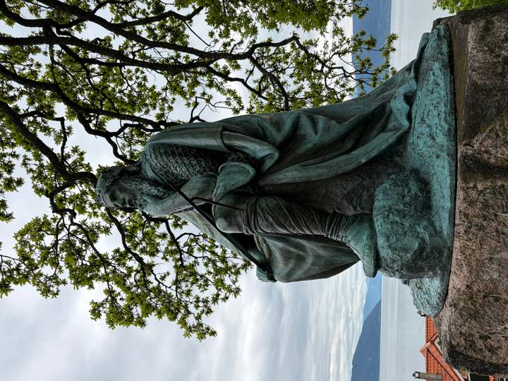 Bronze Viking statue sits pensively beneath leafy branches overlooking a fjord.