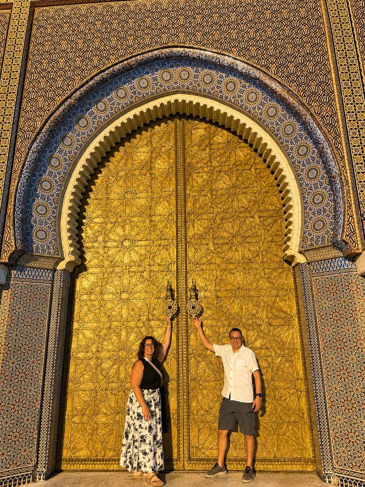 Two tourists raise their hands while standing before the ornate golden doors of the Royal Palace in Fes.