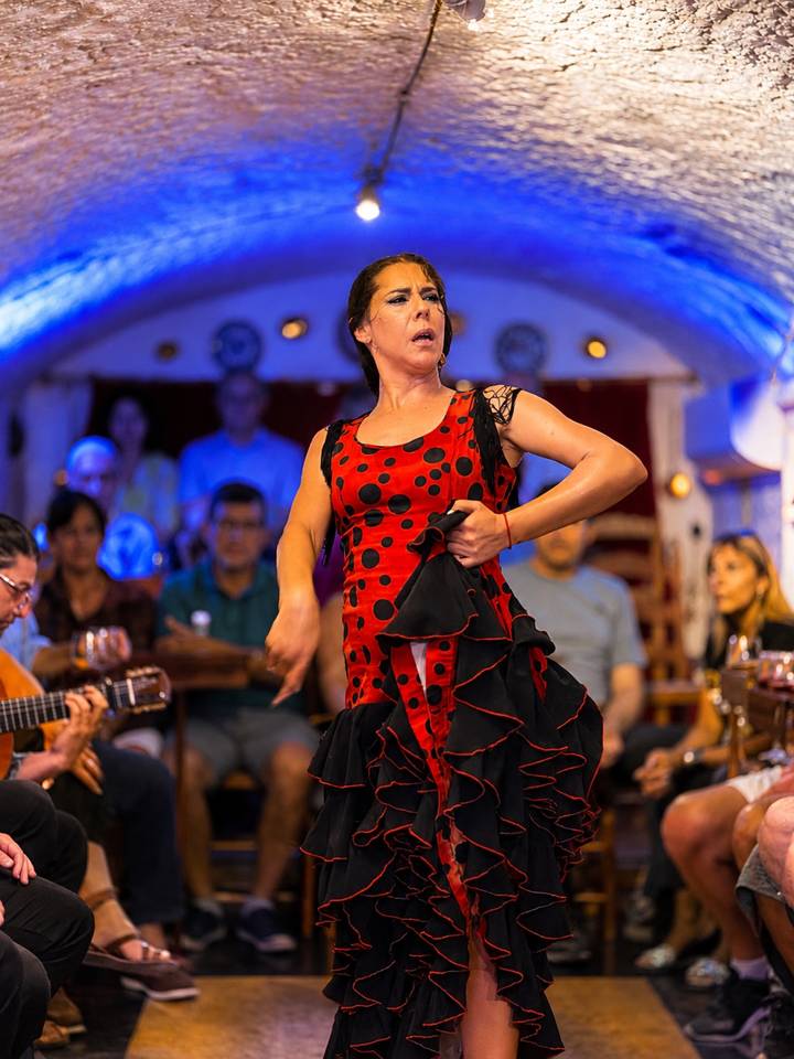 Flamenco dancer in a red and black polka-dot dress performs passionately inside an intimate tablao with musicians and audience.
