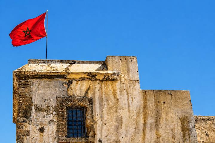 Red Moroccan flag flutters atop an aged stone fortress wall against a vivid blue sky.