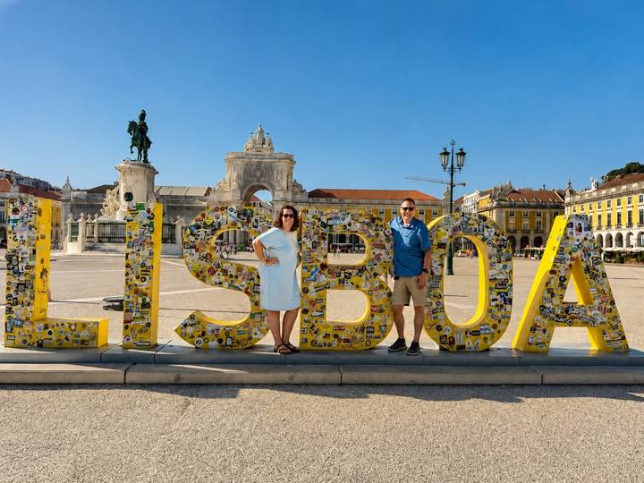 Two tourists pose in front of the large yellow 'LISBOA' sign at Lisbon's Praça do Comércio on a bright sunny day.