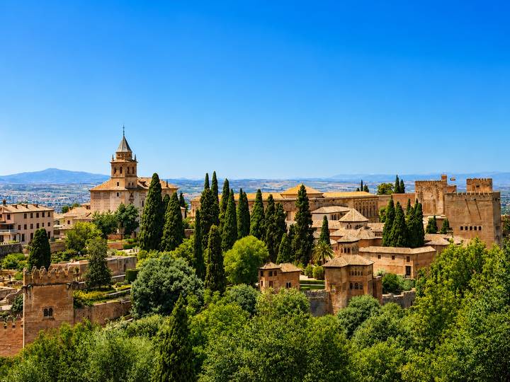 Elevated view of the Alhambra palace complex surrounded by cypress trees with mountains faintly visible in the distance.