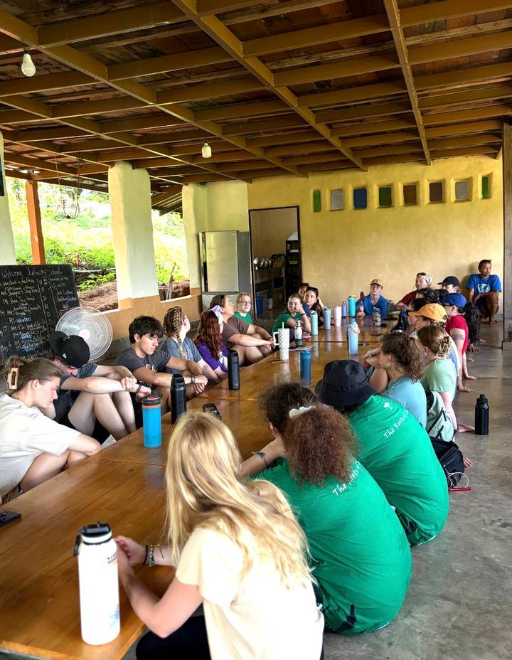Participants sit around a long wooden table inside a rustic meeting hall during a briefing.
