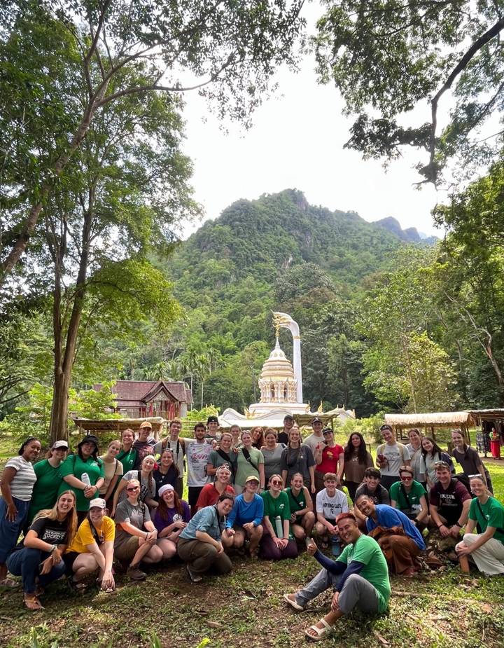 Large group poses before a white and gold temple spire set in lush mountain forest.