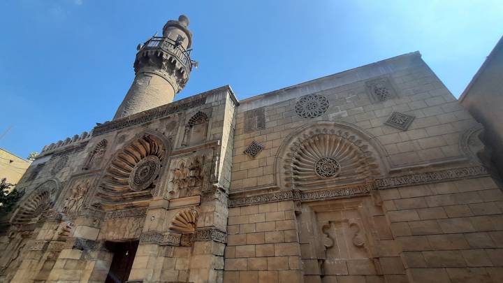 Historic sandstone mosque façade with ornate carvings and a tall minaret under blue sky.