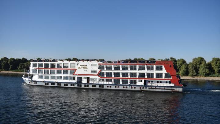 White-and-red Nile cruise ship sailing along calm river against clear blue sky.