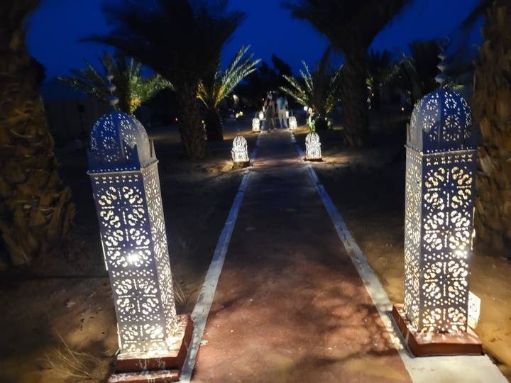 Lantern-lit pathway flanked by palm trees leading to desert tents at night.