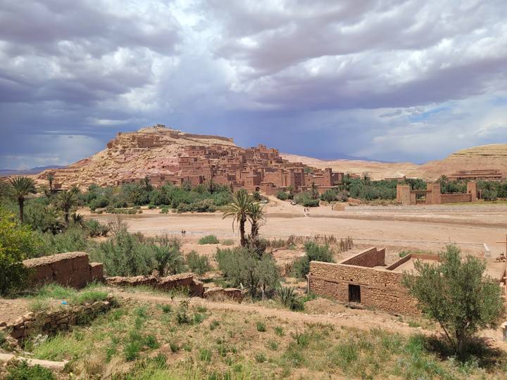 Panoramic view of the fortified clay village of Ait Benhaddou surrounded by palm trees and desert hills under a dramatic sky.