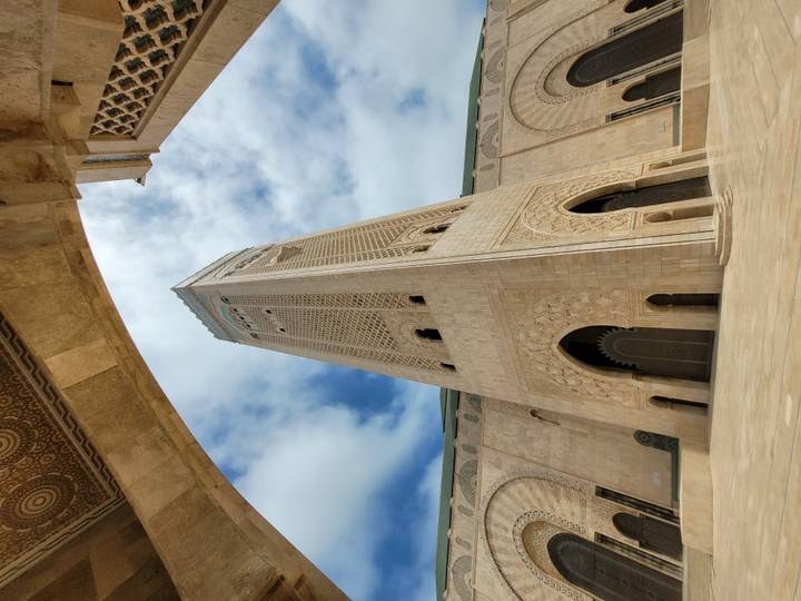 Upward perspective of the ornate minaret and archways of Hassan II Mosque against a partly cloudy blue sky.
