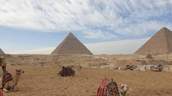 Iconic pyramids of Giza rising from sandy desert with camels and blankets in the foreground under streaked sky.