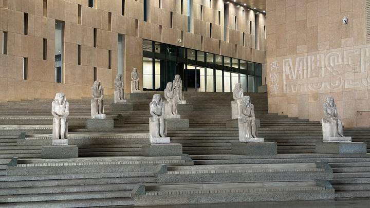 Series of seated ancient statues lining modern stone steps outside a contemporary museum building.