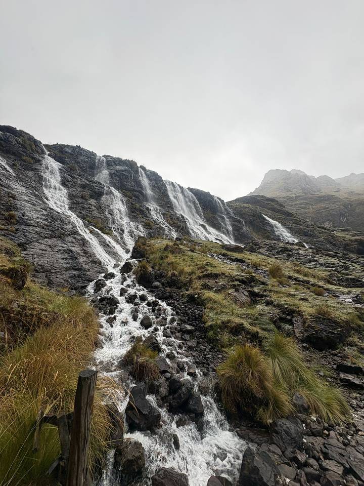Multiple streams of water tumble down a steep, rocky Andean cliff under misty grey skies.