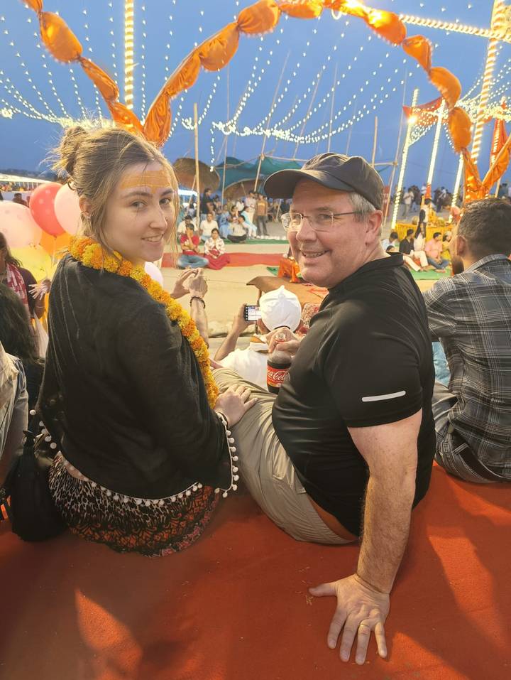Two travelers wearing marigold garlands look back while seated among a festive crowd during an Indian ceremony.