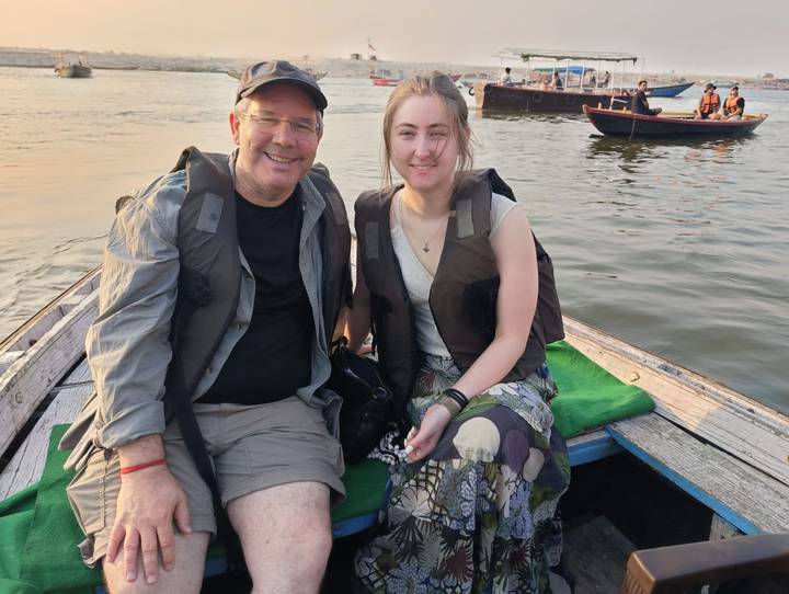 A man and woman sit smiling on a small wooden boat on the Ganges River during golden evening light.