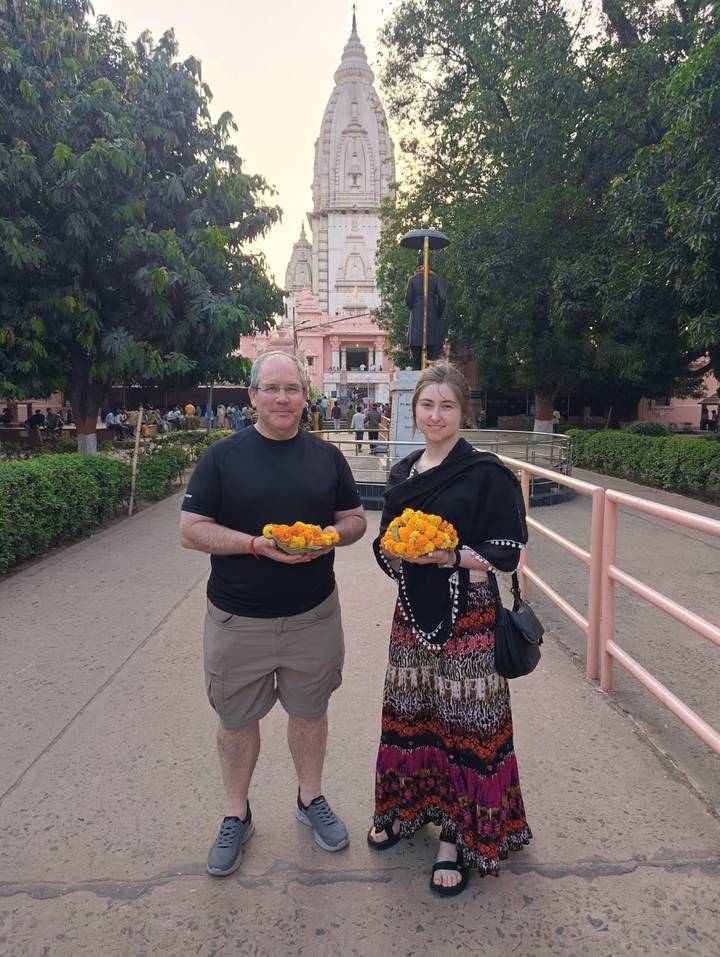 Travelers hold baskets of marigold flowers outside a pink temple complex surrounded by trees and visitors.