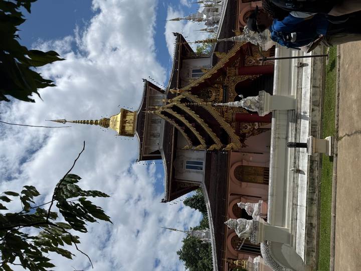 An ornate Thai temple with layered golden roof tiers and a shining spire rises against a partly cloudy sky.