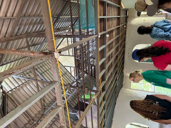 A group of visitors listen and look upward inside a rustic building with exposed beams and metal roof.