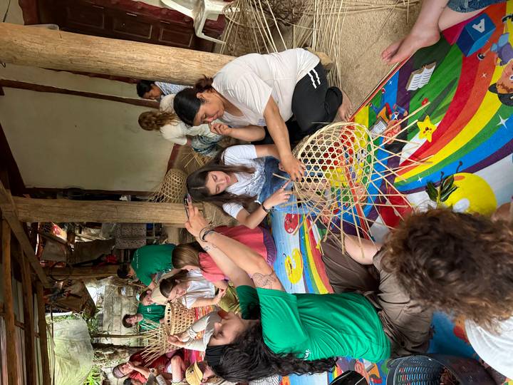 Travellers sit on colourful mats learning traditional bamboo weaving from local women in an open wooden shelter.
