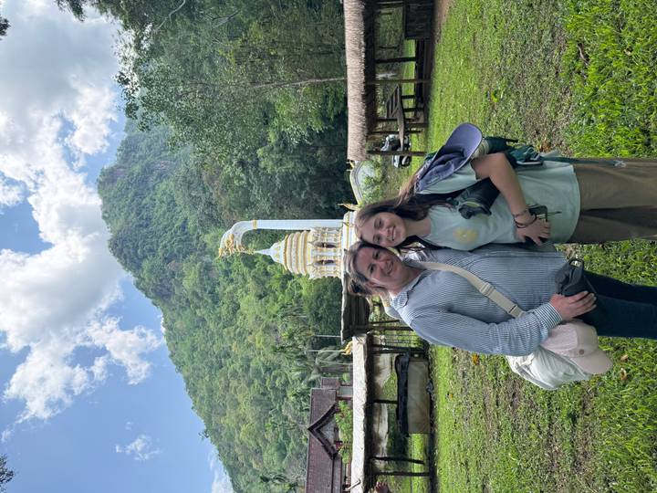 Two women stand smiling in a green valley with a white stupa and forested mountain rising behind.