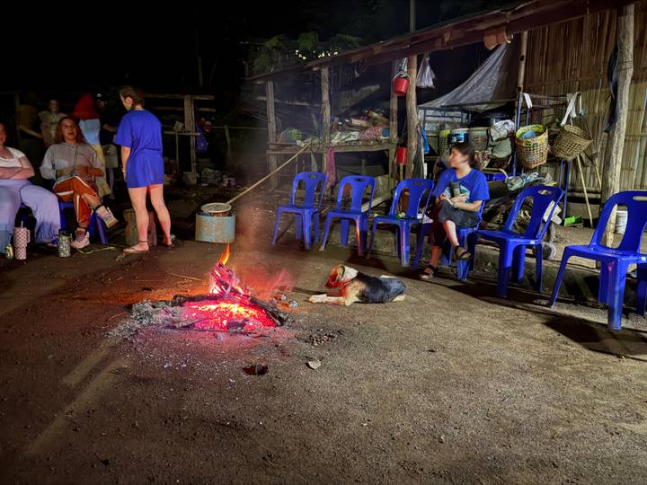 Travellers gather around a glowing campfire at night while a dog lies nearby and blue chairs ring the space.