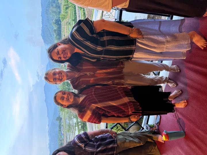 Three women dressed in woven tops smile together on a patio with rice fields and mountains behind.