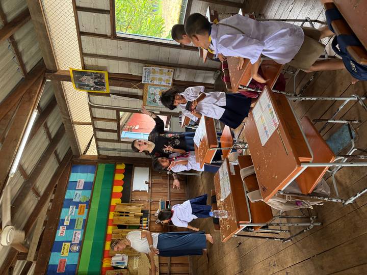 School children in uniform engage in lively activity inside a wooden classroom with volunteers.