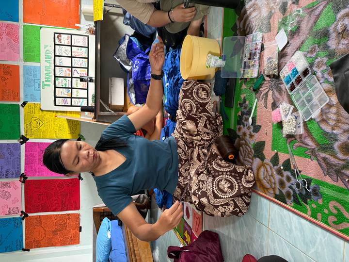 A woman sits cross-legged on a patterned mat organising beads for handicrafts in a colourful room.