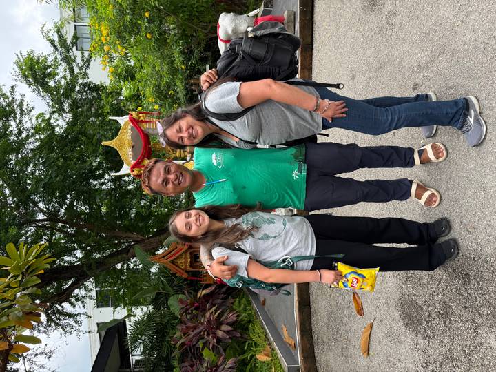 Travellers pose with their smiling guide outside a small shrine surrounded by tropical plants.